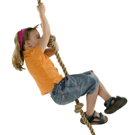 A young girl with blonde hair in a pink headband, wearing an orange t-shirt and denim shorts, climbing a children’s knotted climbing rope against a solid white background.
