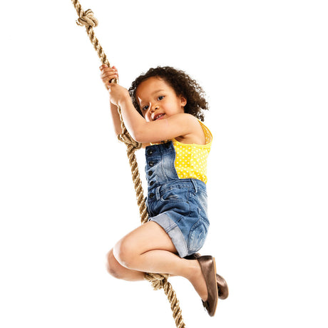 A child with curly hair climbing a knotted climbing rope against a solid white background, wearing a yellow patterned tank top, denim overalls, and brown shoes.