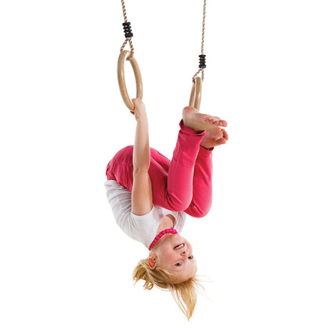 A young girl hanging upside down from wooden gymnastic rings on a white background, wearing a white t-shirt, pink leggings, and a pink headband.
